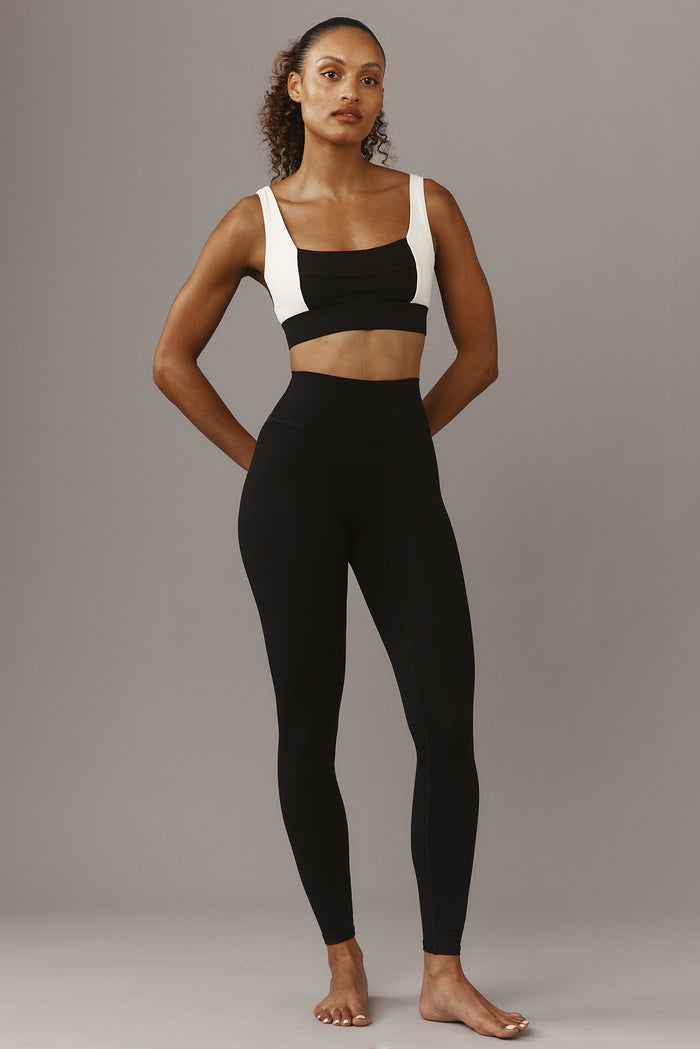 Athletic woman standing barefoot against a gray backdrop wearing black high-waist leggings and a black-and-white sports bra.