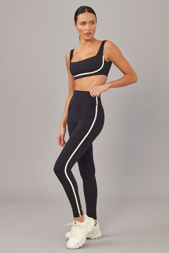 Three-quarter view of Acadia Legging 27-inch in Black/Ivory showcasing high waist and ivory side stripe on model against gray background