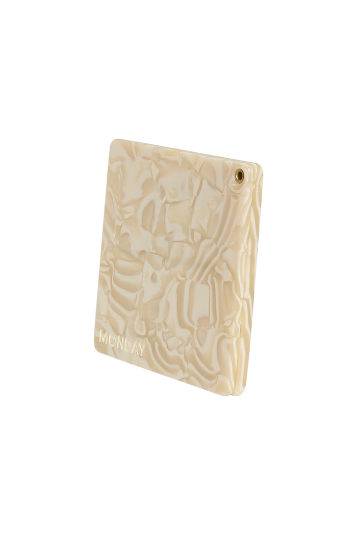 Angled view of Acklins Mirror (Ivory Swirl) showing cream marbled acrylic face, embossed MONDAY logo and brass grommet on white background