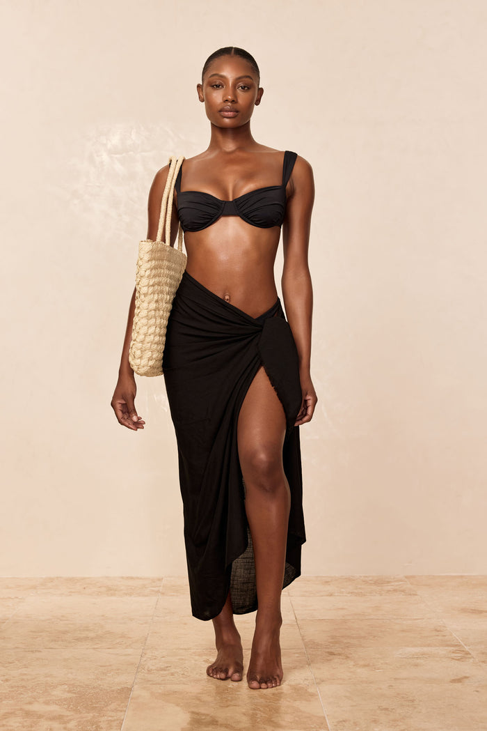 Front view of Amalfi Sarong in black on model showcasing wrap knot, raw edge hem and thigh-high side slit against neutral beige backdrop