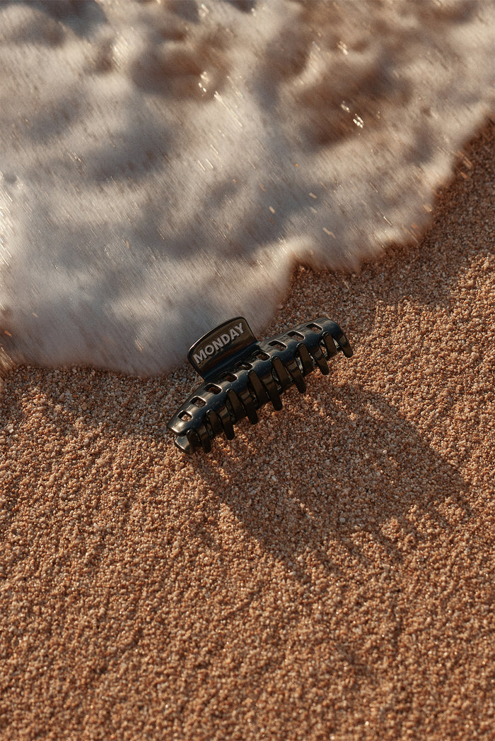 Black hair claw clip labeled MONDAY resting on wet sand as a wave's foam approaches