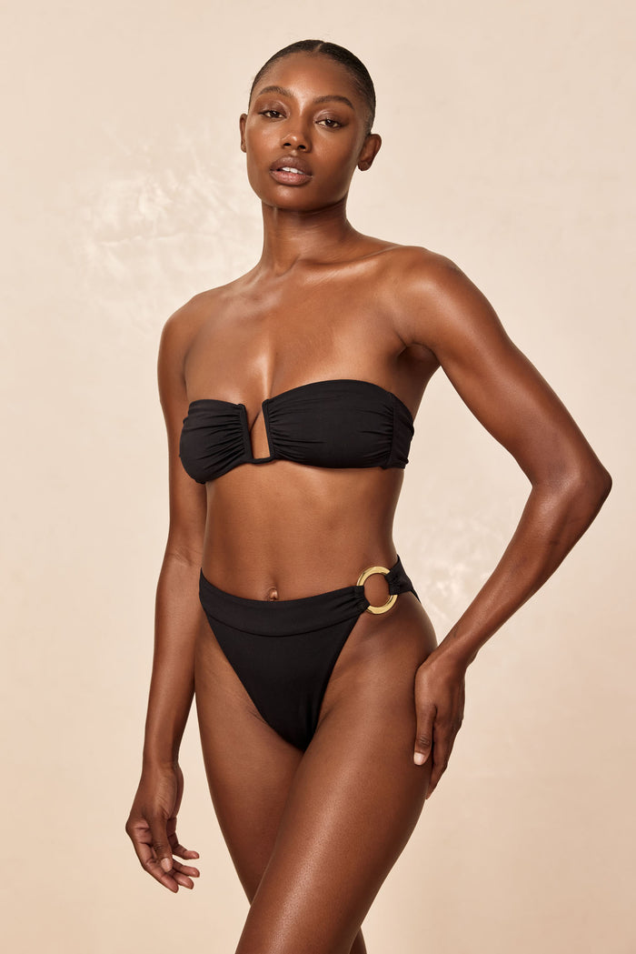 Three-quarter front of Antigua Bottom in black showcasing high-waisted fit, high-cut leg and chunky gold ring on left hip on beige background