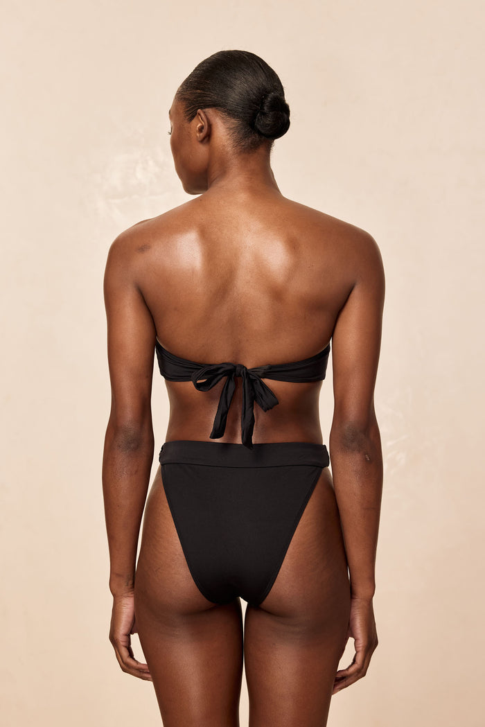 Back view of Antigua Bottom in black on model, showcasing high-waisted high-cut silhouette and bandeau tie on beige background