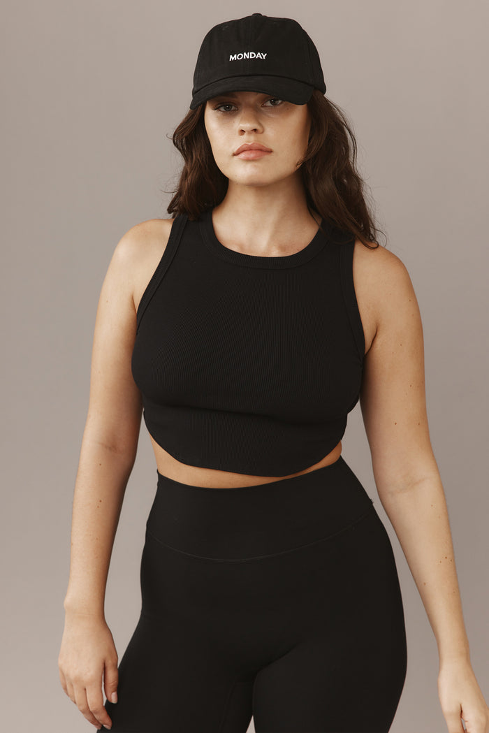 Woman wearing a black ribbed cropped tank and high-waist leggings, black baseball cap embroidered "MONDAY".