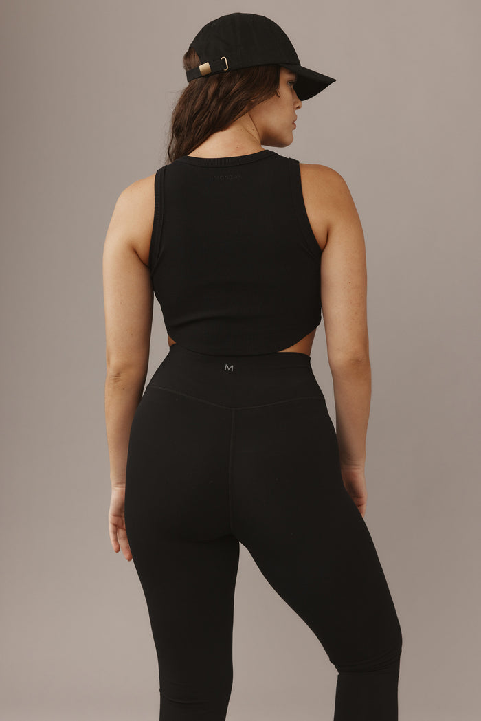 Back view of woman wearing black ribbed tank ('MONDAY' at neckline), high-waist black leggings with 'M' logo, and black cap
