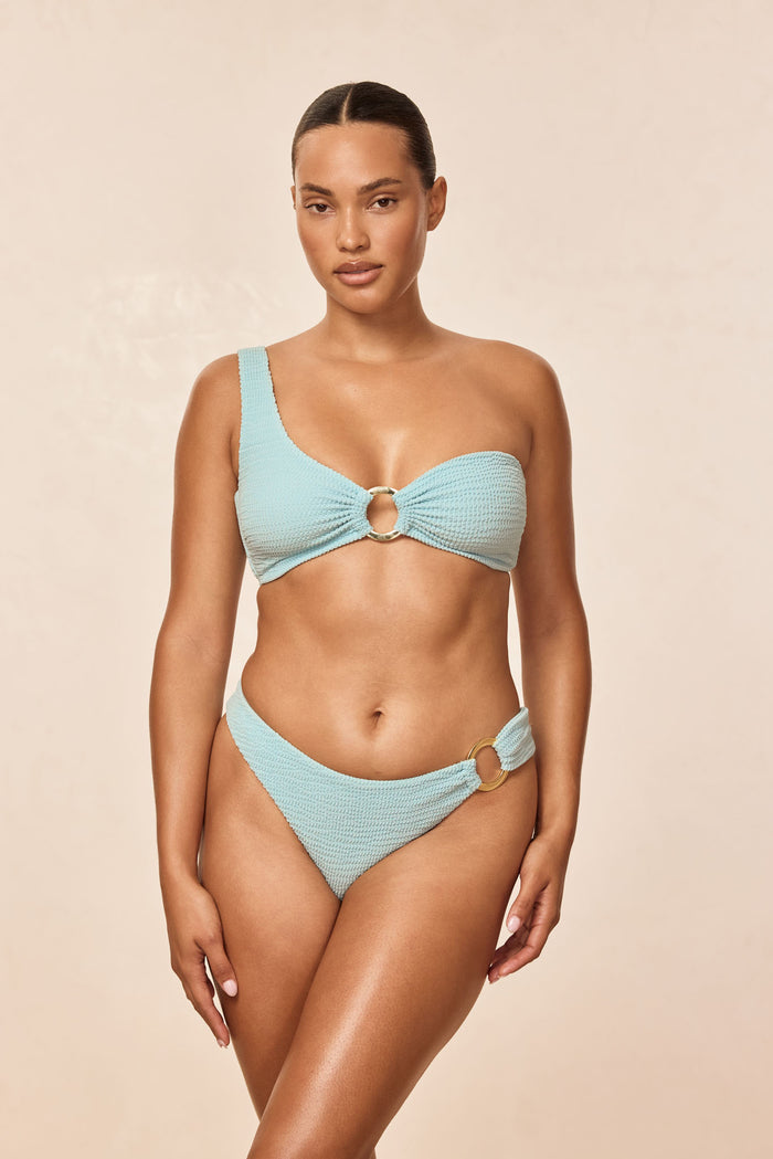 Front view of Antigua Top - Light Slate Crinkle showcasing one-shoulder strap, textured crinkle fabric and center gold ring on a beige background