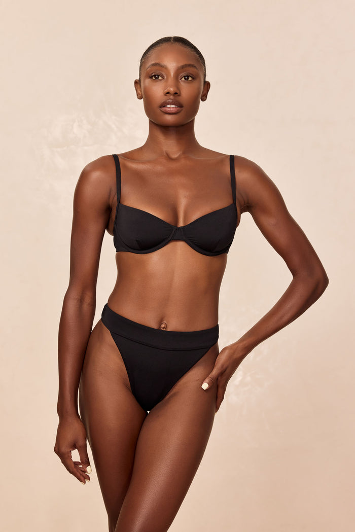 Front view of Argentina Bottom in black on model showcasing high-cut leg and fold-over waistband against beige background