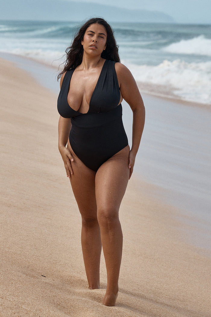 Front view of Aruba One Piece in black showcasing deep V neckline, wide shoulder straps, and supportive waist panel on a sandy beach