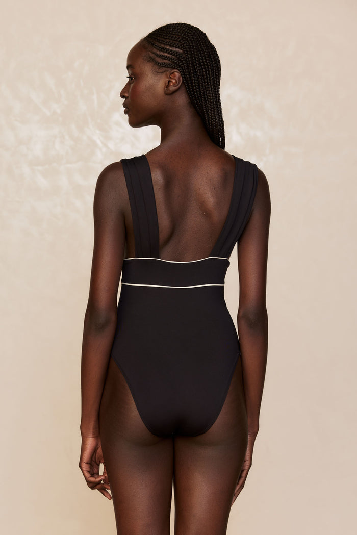 Back view of Aruba One Piece in Black/Ivory showcasing wide shoulder straps and ivory piping at the waist with beige background