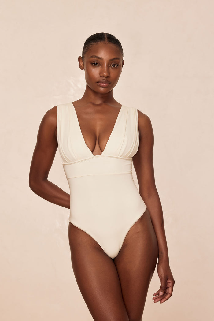 Front view of Aruba one-piece swimsuit in ivory with V-neck, ruched wide straps and high-cut leg on beige background
