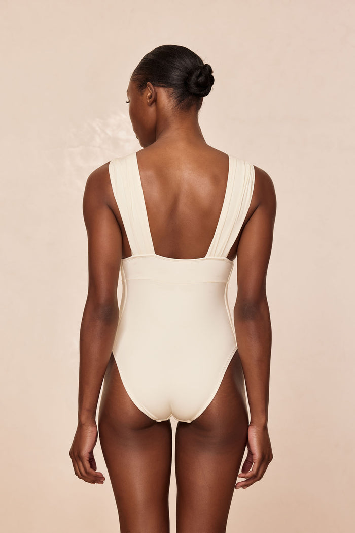 Rear view of Aruba One Piece swimsuit in ivory with low square back, wide pleated straps, moderate coverage on beige backdrop