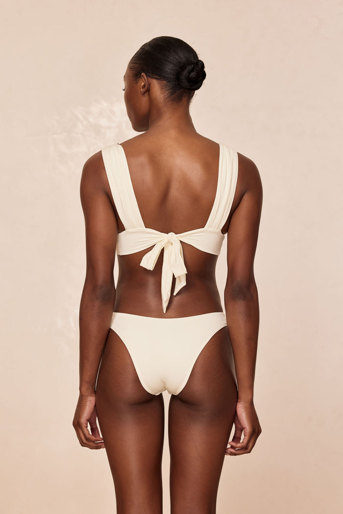 Back view of Aruba Top in Ivory on model showing wide pleated shoulder straps and tied adjustable back closure against a neutral background