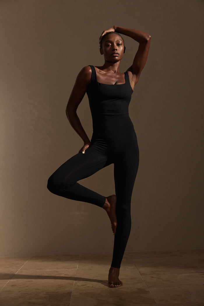 Front view of Aspen Catsuit in black jersey showcasing square neckline, high waist seam and tapered full-length legs on warm brown studio backdrop
