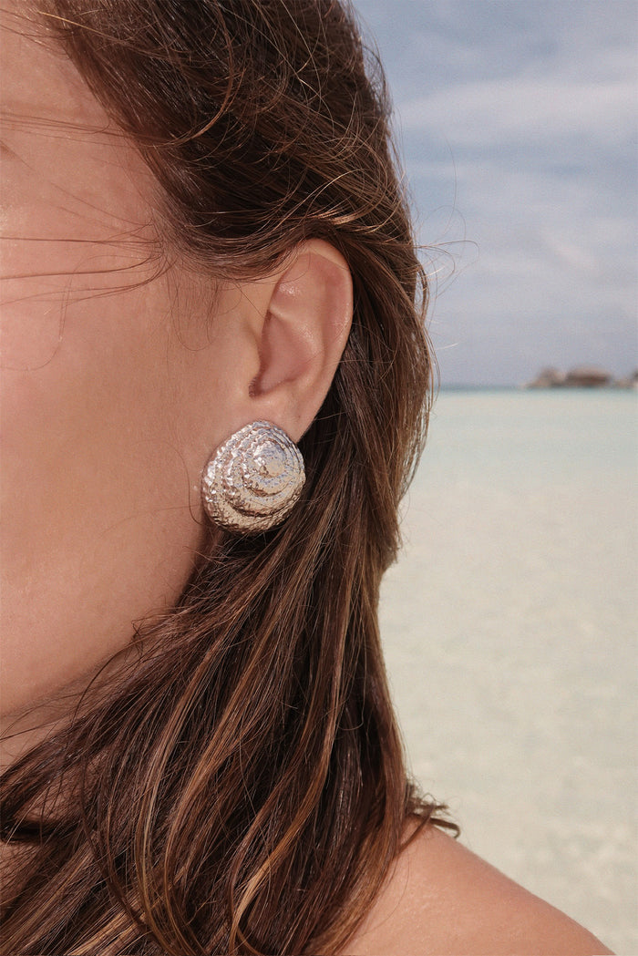 Close-up of Barbados silver earrings on model’s ear showcasing hammered spiral texture with blurred beach and sea background