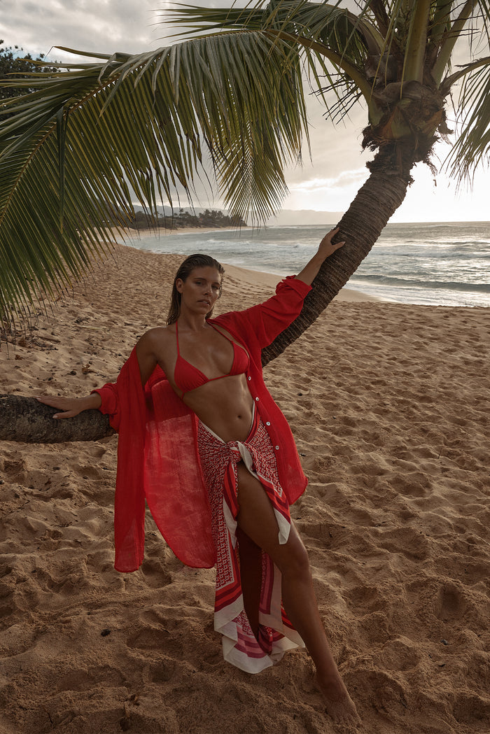 Model wearing Palma Top in Chili Pepper with matching sheer tunic leaning on a palm tree on a sandy beach