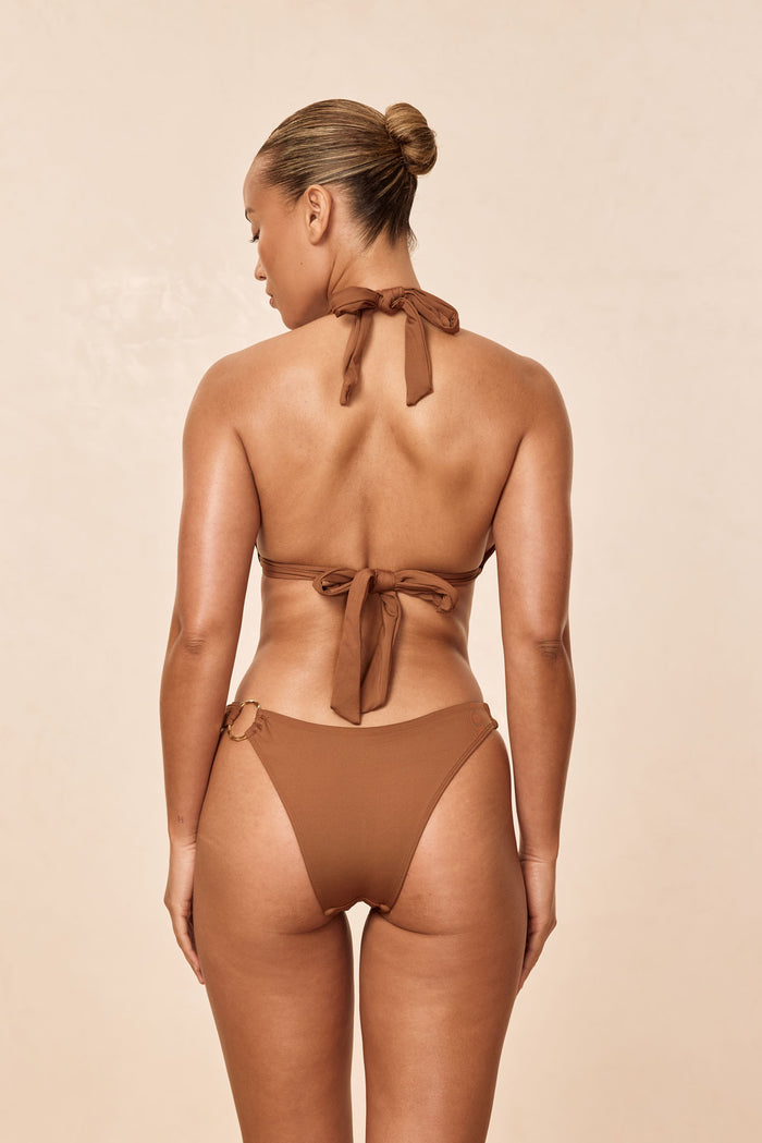Back view of Bari Bottom in Caramel showing high-cut leg, mid-rise, cheeky coverage and organic gold ring on beige background