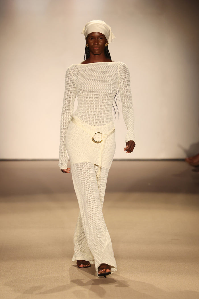 Model walking the runway in an ivory lace crochet long-sleeve knit dress with belted waist and matching headscarf