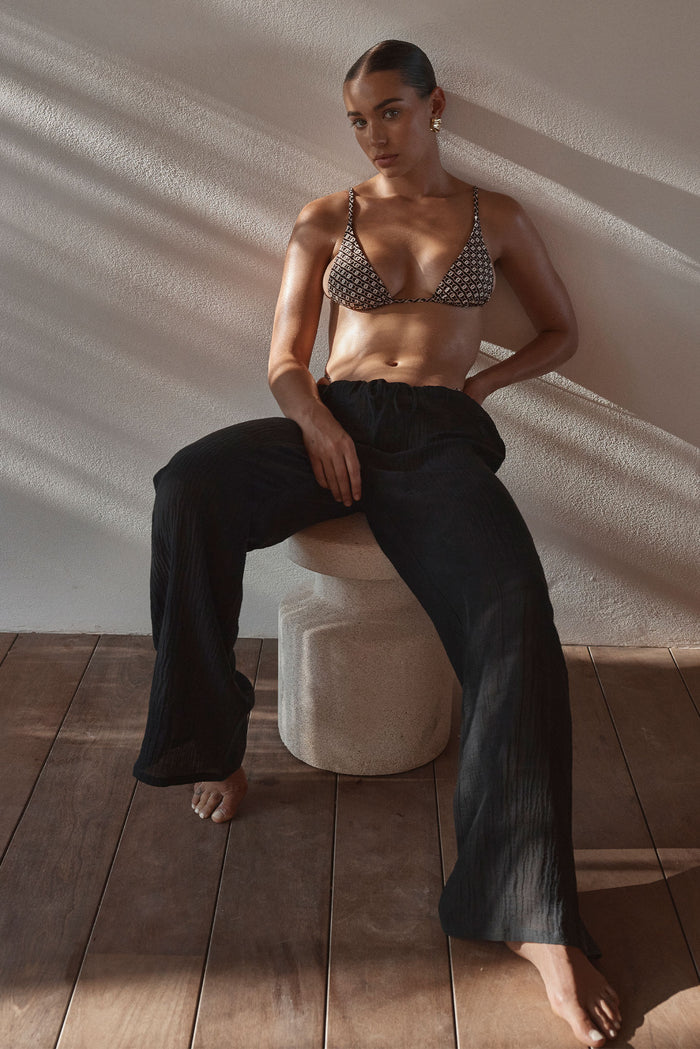 Seated front view of Belize Pant in Black Crinkle Linen showing relaxed wide leg fit and drawstring waist with sunlit wall