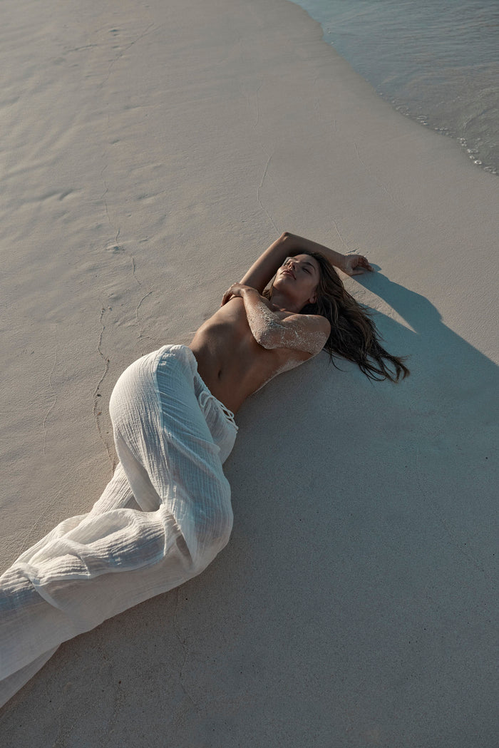 Reclining view of Belize Pant in Ivory Crinkle Linen showcasing sheer crinkled fabric and relaxed wide-leg fit on white sand