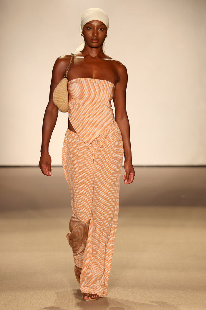 Model on runway wearing peach Micro Geo Silk Belize pants and matching strapless top with cream headscarf and woven bag