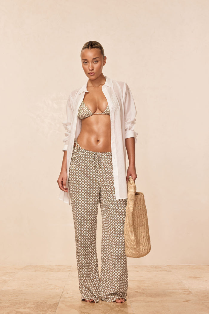 Model wearing Tile Geo Belize pants and matching triangle bikini top with an open white shirt, holding a straw tote against a beige background