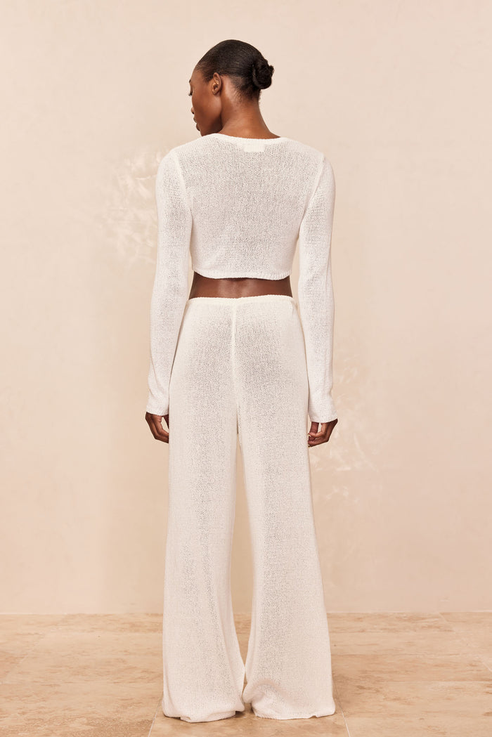 Back view of model wearing white sheer knit long-sleeve cropped top and high-waist wide-leg knit pants on beige backdrop
