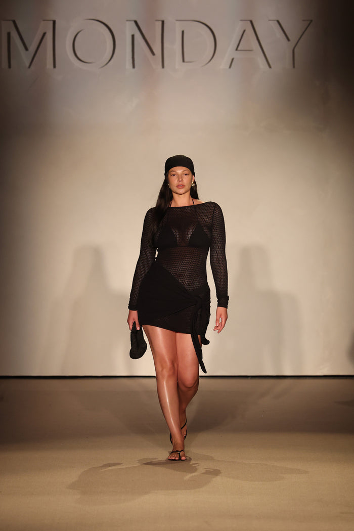 Front view of Bodrum Mini Dress Black Lace Crochet showcasing sheer long-sleeve crochet knit and wrap-tie mini skirt with runway backdrop