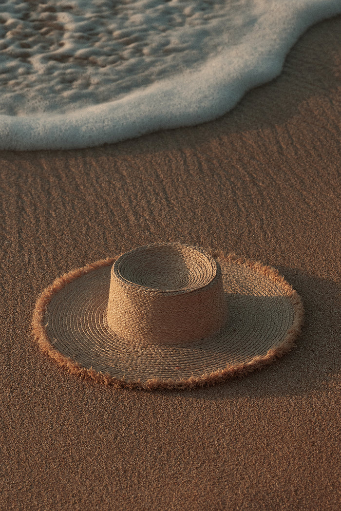 Overhead view of Bondi Hat small brim straw hat showcasing frayed brim and flat crown on wet sand by a foamy wave