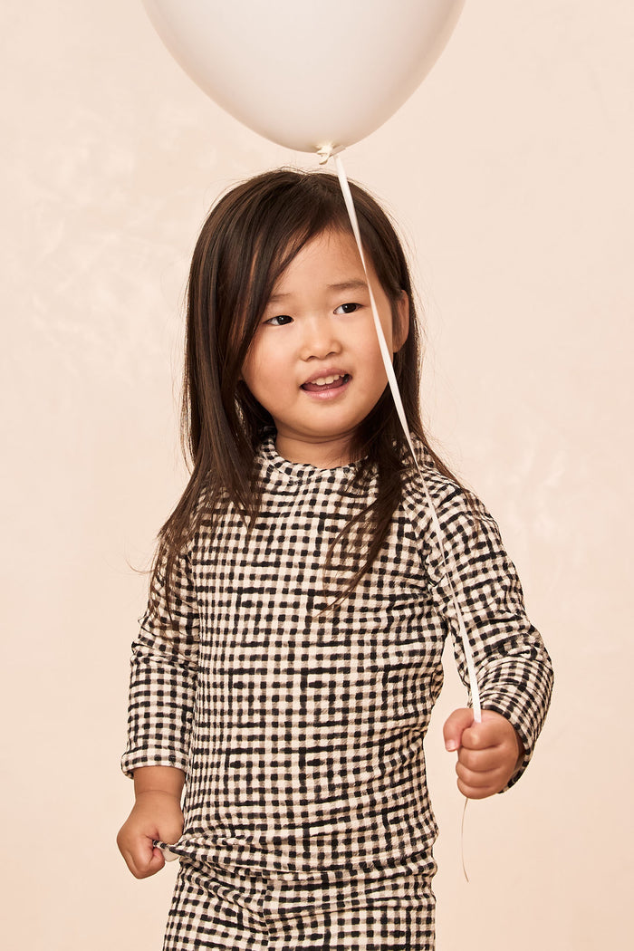 Front view of Bondi Rashguard Kids in black gingham on child model, fitted long-sleeve mock-neck rashguard with pale beige background