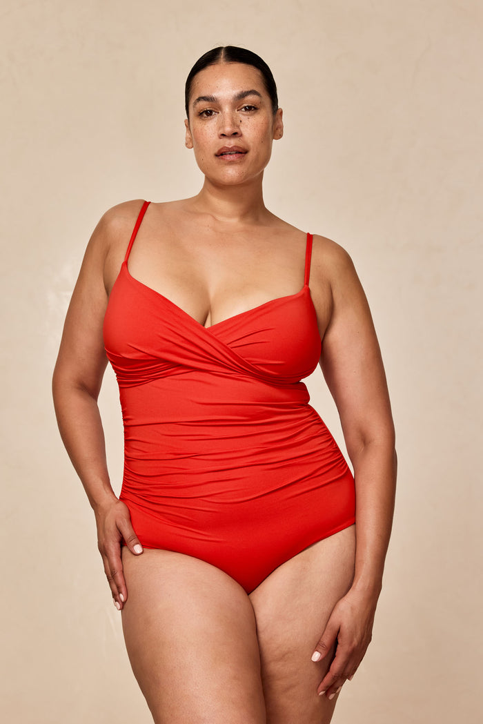 Woman wearing a red one-piece swimsuit standing with hands near hips against a neutral beige background.
