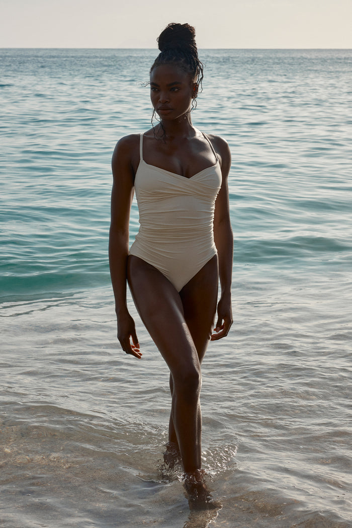 Three-quarter front of Bordeaux One Piece full coverage in Ivory showcasing ruched bodice and thin straps with sunlit sea background
