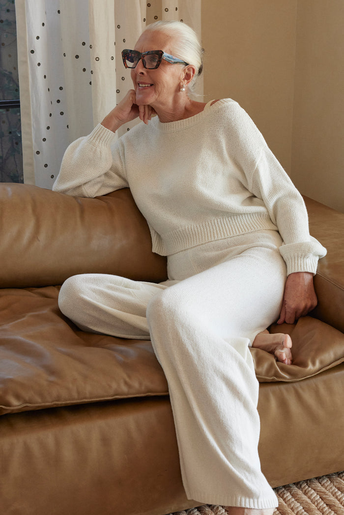 Seated view of Brampton Island sweater in white showing cropped ribbed hem and relaxed sleeves with matching pants on tan leather sofa