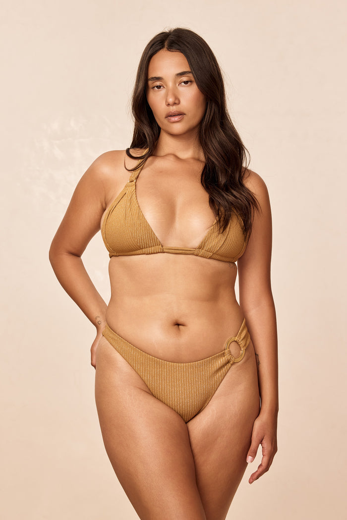 Front view of Bronte Bottom in antique gold lurex rib showing high-cut ribbed silhouette with hip ring on beige background