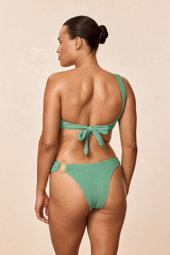 Back view of Bronte Bottom in Pond Crinkle showcasing high-cut cheeky coverage and gold hip ring on beige background