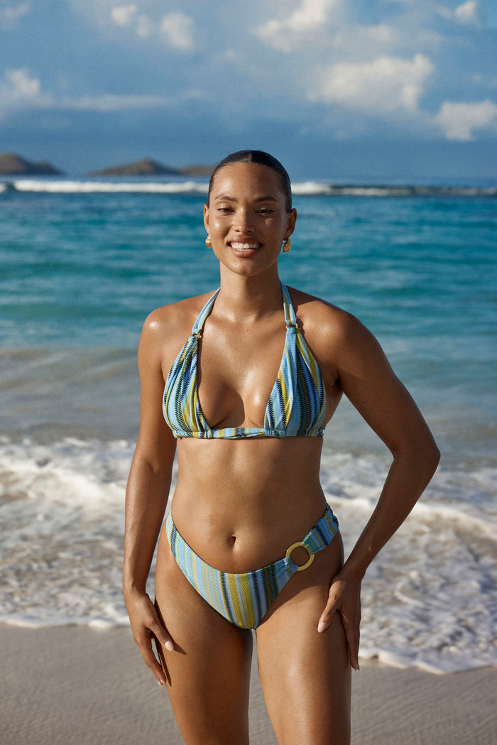 Front view of Bronte Bottom in Sardinia Stripe Azure showcasing mid-rise cut, gold side ring, and striped textured fabric on a beach