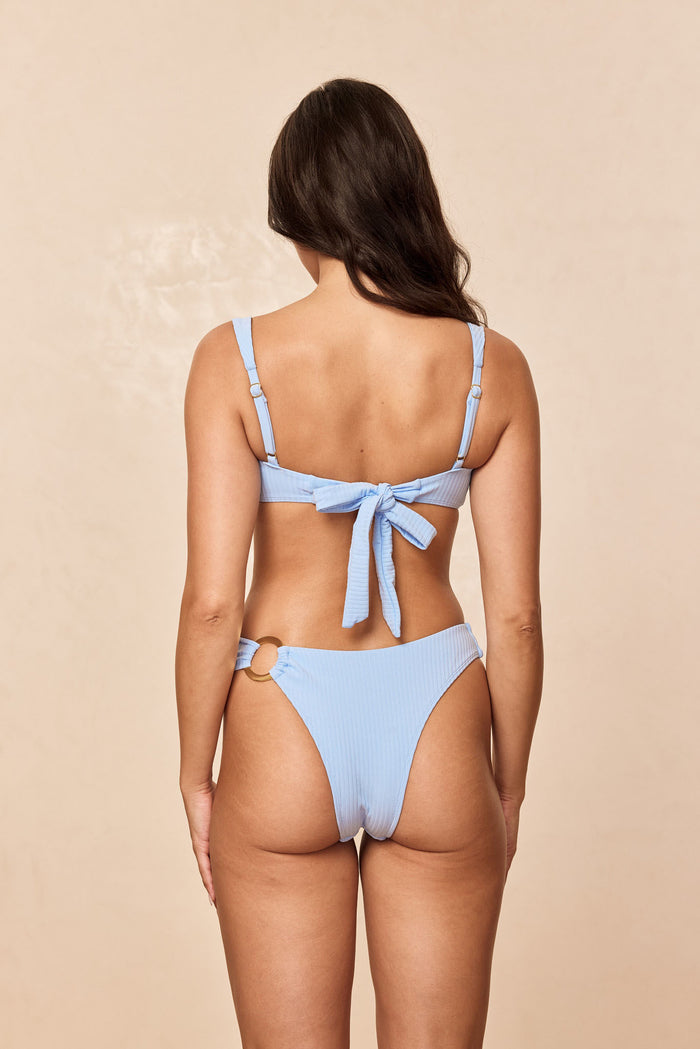 Back view of Barbuda Top in Sky Rib with tie-back closure and adjustable straps on model against beige background