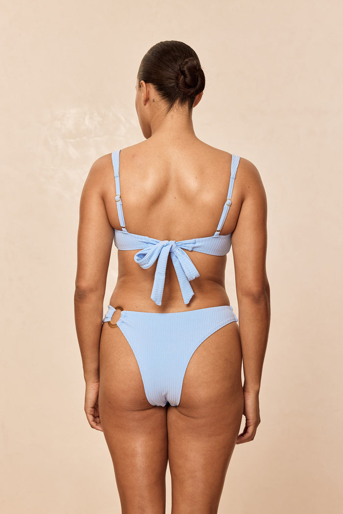 Rear view of Barbuda Top in Sky Rib on model showcasing tie-back bow, adjustable straps, and ribbed texture on beige background