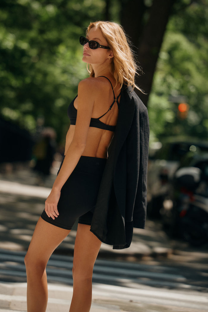 Rear three-quarter view of Brooklyn Bra in black showcasing triangle cup shape and thin adjustable cross-back straps on a sunlit city sidewalk