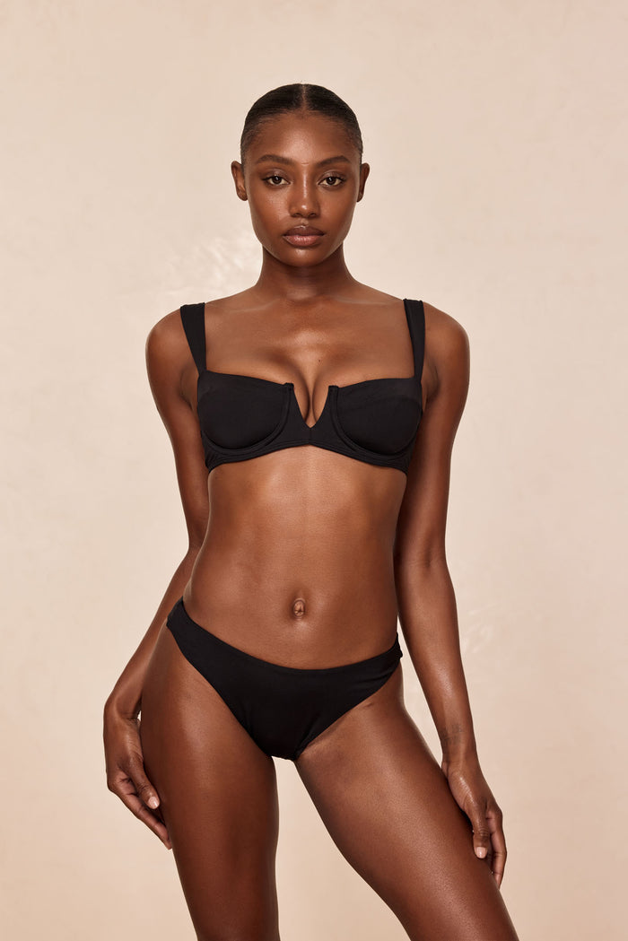Front view of Byron Bottom in black, mid rise bikini bottom on model showing moderate coverage with beige studio background