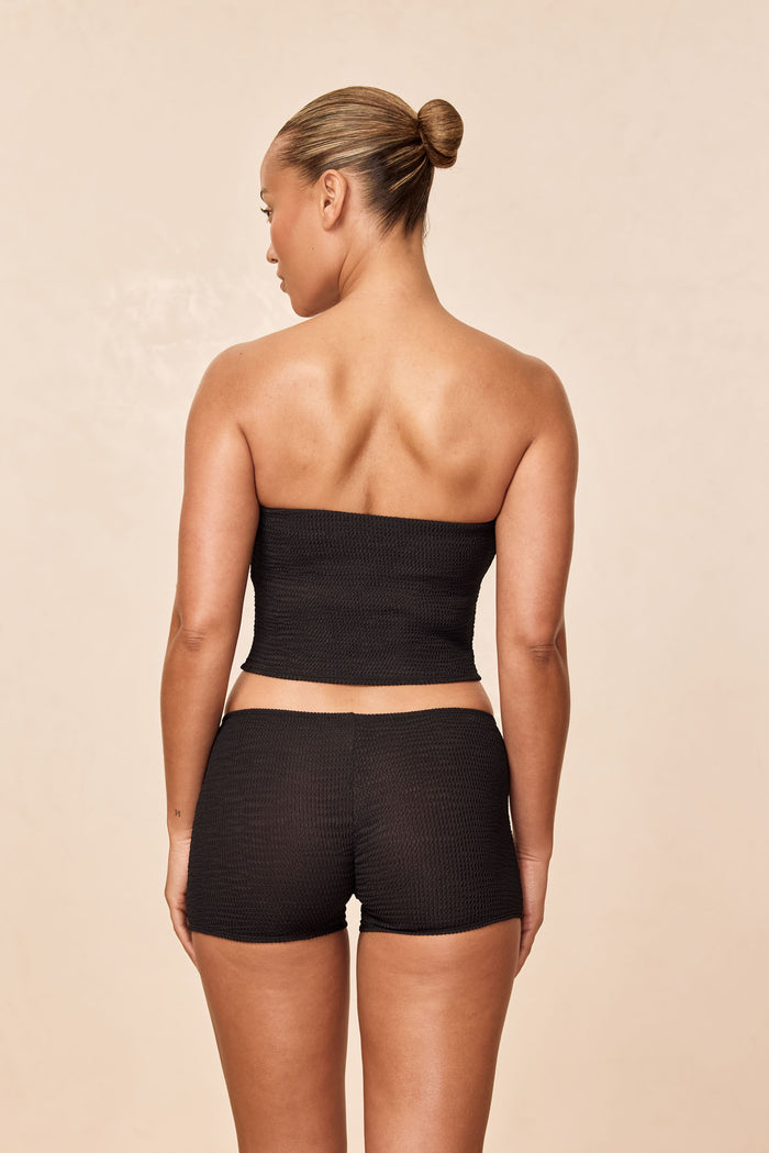 Back view of Cali Short in Black Crinkle showcasing high-waist textured knit boyshort fit on model against beige backdrop