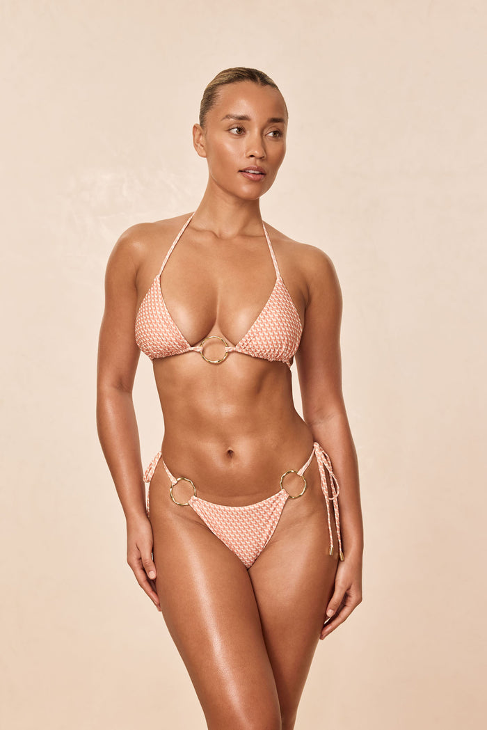 Front view of Cannes bottom in peach Micro Geo on model showing side-tie straps and gold ring hips on beige background
