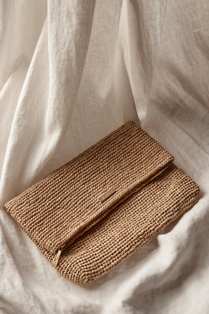 Angled flat lay of Cannes Clutch showing tan woven raffia, fold-over flap with gold logo plaque and visible zipper on draped cream linen
