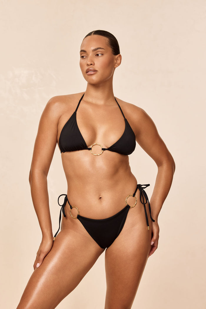 Front view of Cannes Top in black on model, triangle halter with center gold ring and tie straps against beige background