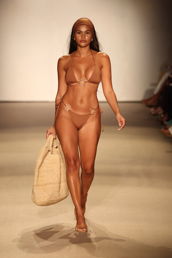 Model walking runway wearing caramel Cannes tie-side bikini bottom with ring hardware and matching top, carrying woven beach bag
