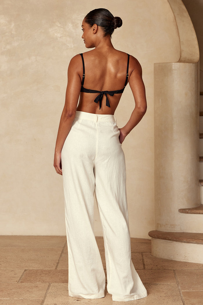 Back view of a woman wearing a black tie-back bralette and high-waist cream wide-leg linen pants against plaster stairs.