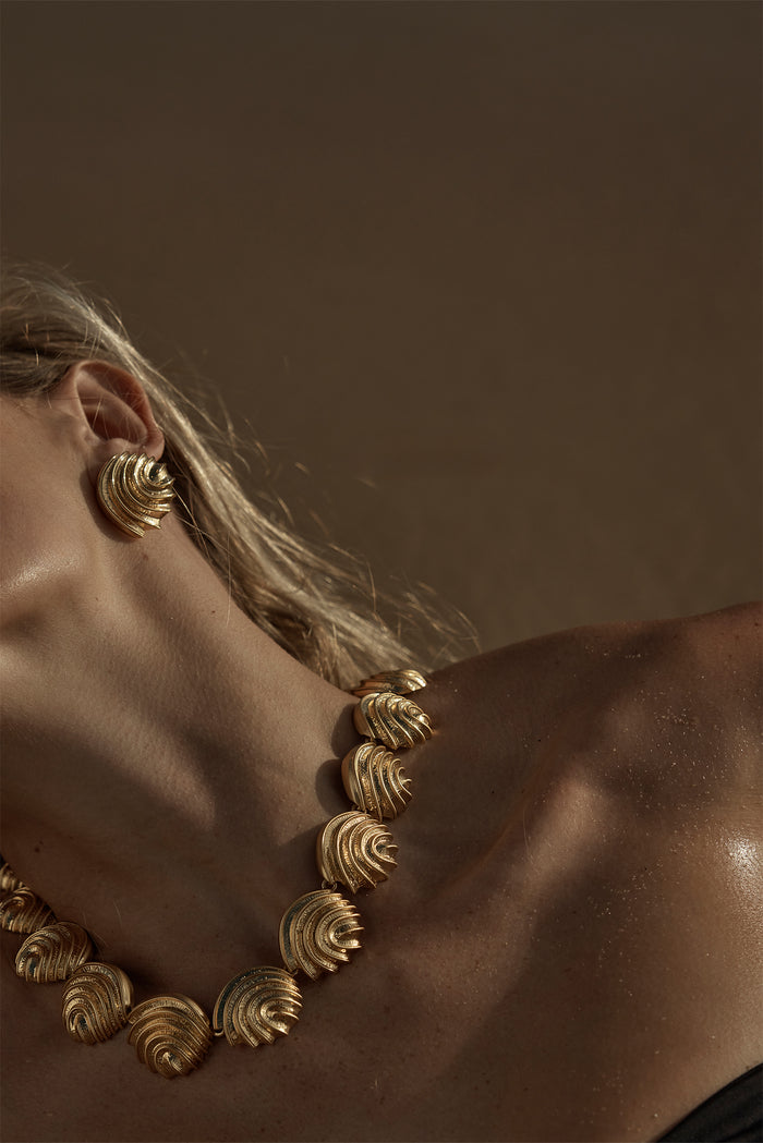 Woman's neckline wearing gold scalloped shell-link necklace and matching earring against warm brown background.