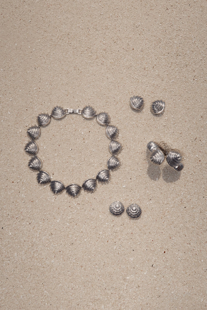Flat lay of Antigua Cuff in silver showing linked seashell-shaped panels and clasp on sand with three pairs of matching shell earrings