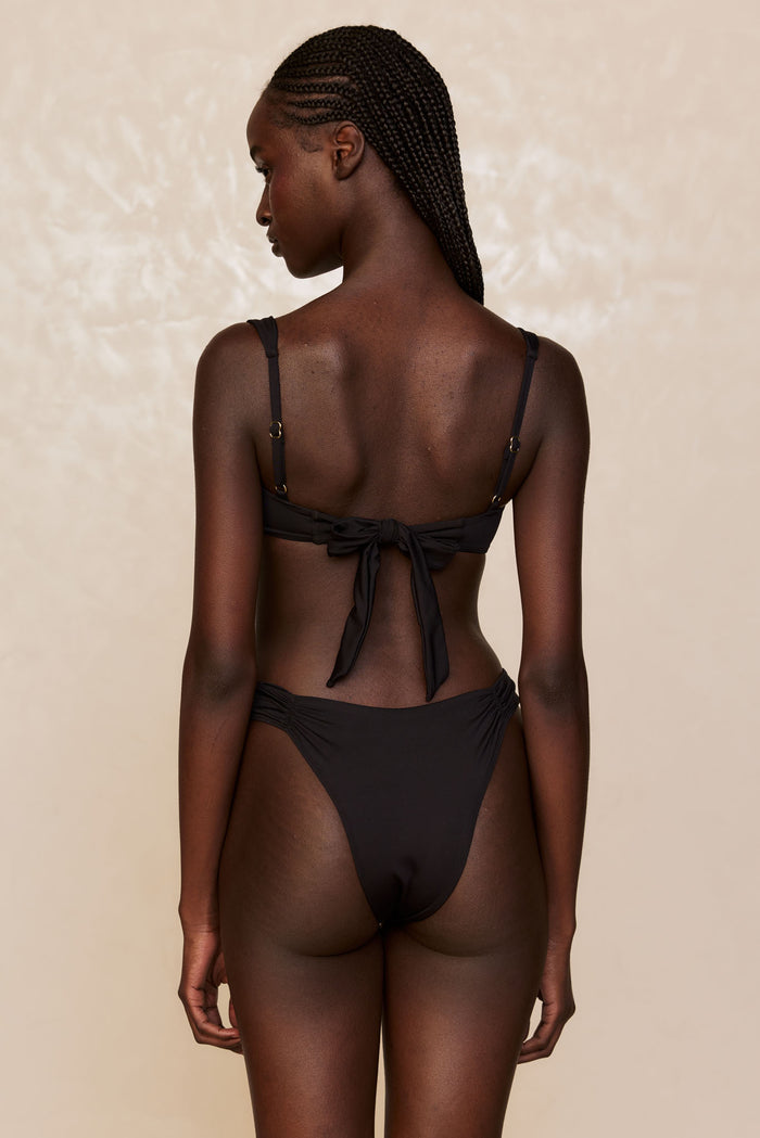 Rear view of Clovelly High Leg Bottom in black showcasing high-cut leg and shirred side hip detail on model against beige background