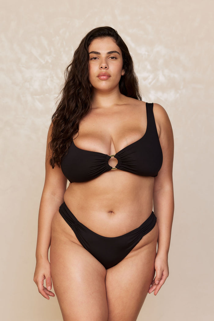 Front view of Clovelly High Leg Bottom in black on a plus-size model showing high-cut leg and shirred side hip detail against a beige backdrop