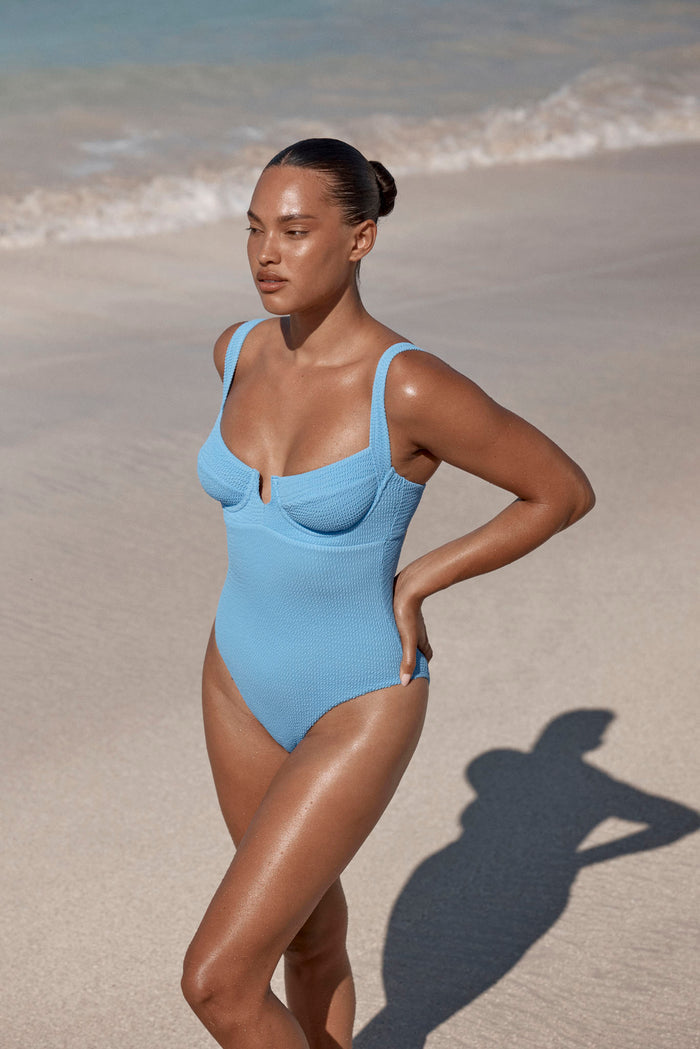 Three-quarter view of Clovelly One Piece in Azure Crinkle showcasing underwire cups and high-cut leg on a sandy beach